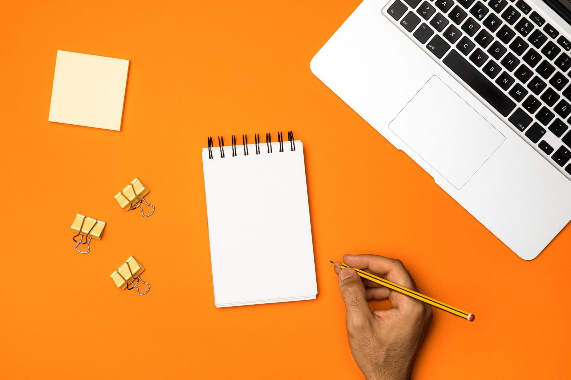Closeup of man holding pencil with notebook, laptop, metal grip clips, and sticky notes.