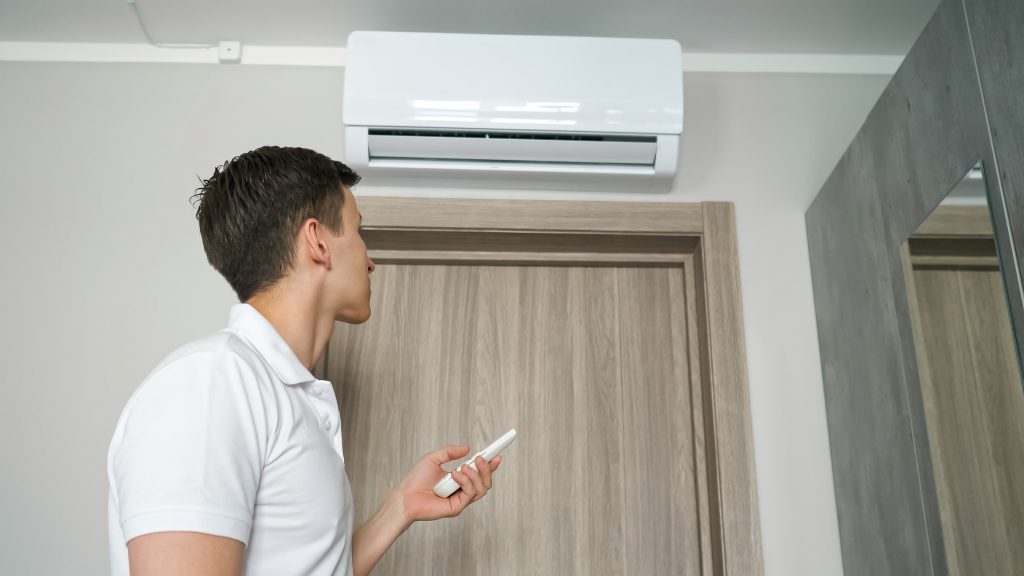 Photo of man looking at AC unit with remote control in hand illustrates blog: "My Air Conditioner Won’t Stay On. What To Do?"